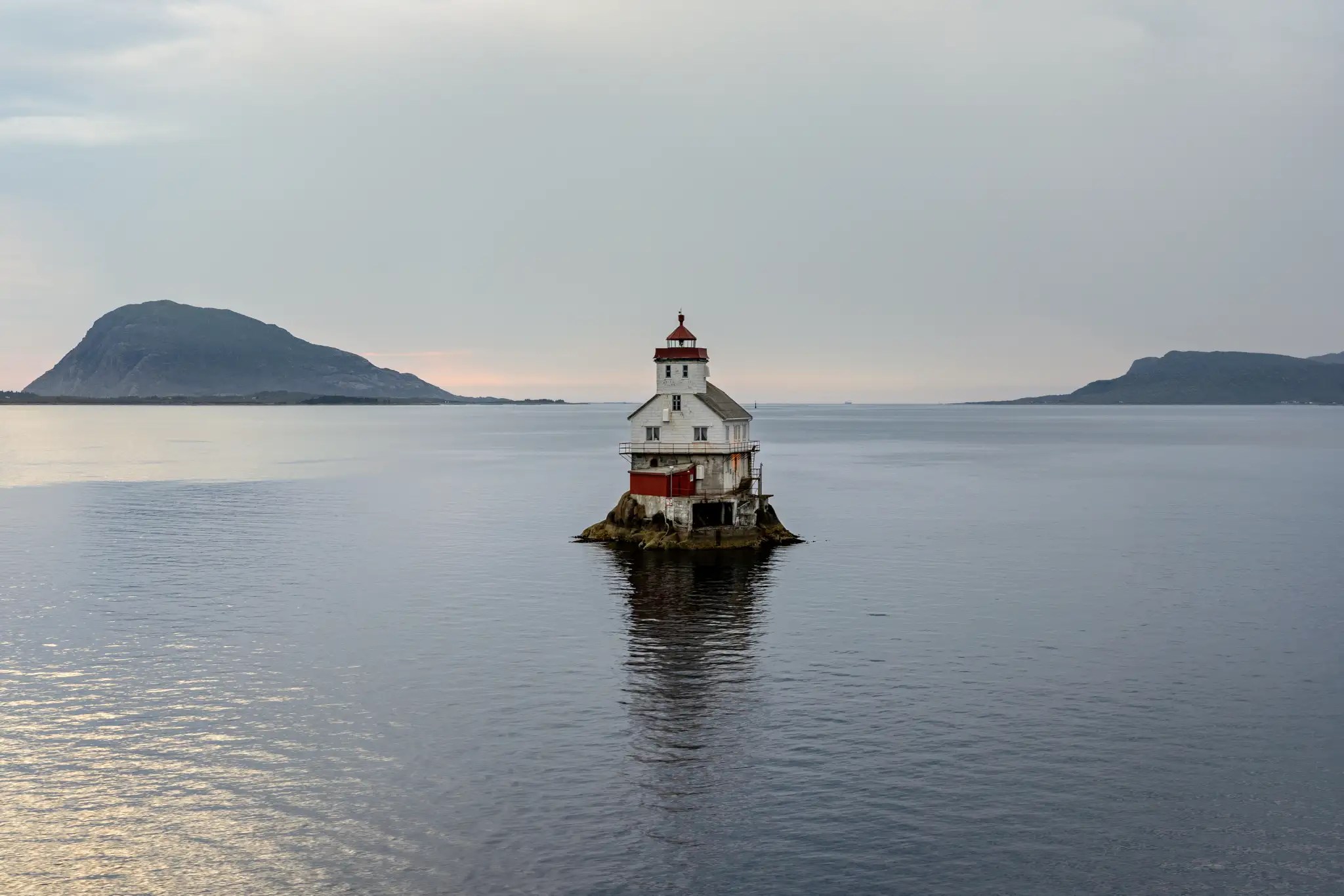 Leuchtturm auf kleinem Felsen im ruhigen Meer, im Hintergrund zwei bewaldete Inseln unter bewölktem Himmel