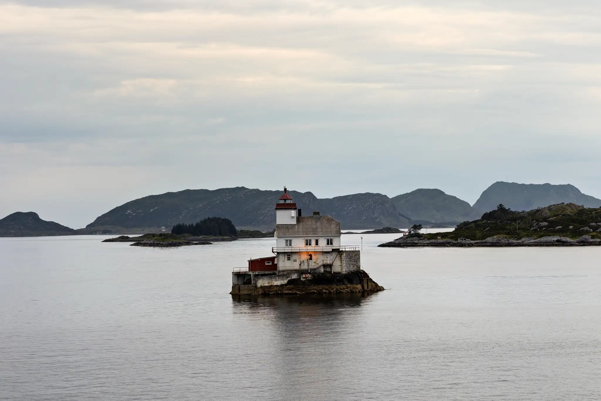Leuchtturm auf kleinem Felsen im ruhigen Meer vor bewölktem Himmel und Inseln im Hintergrund