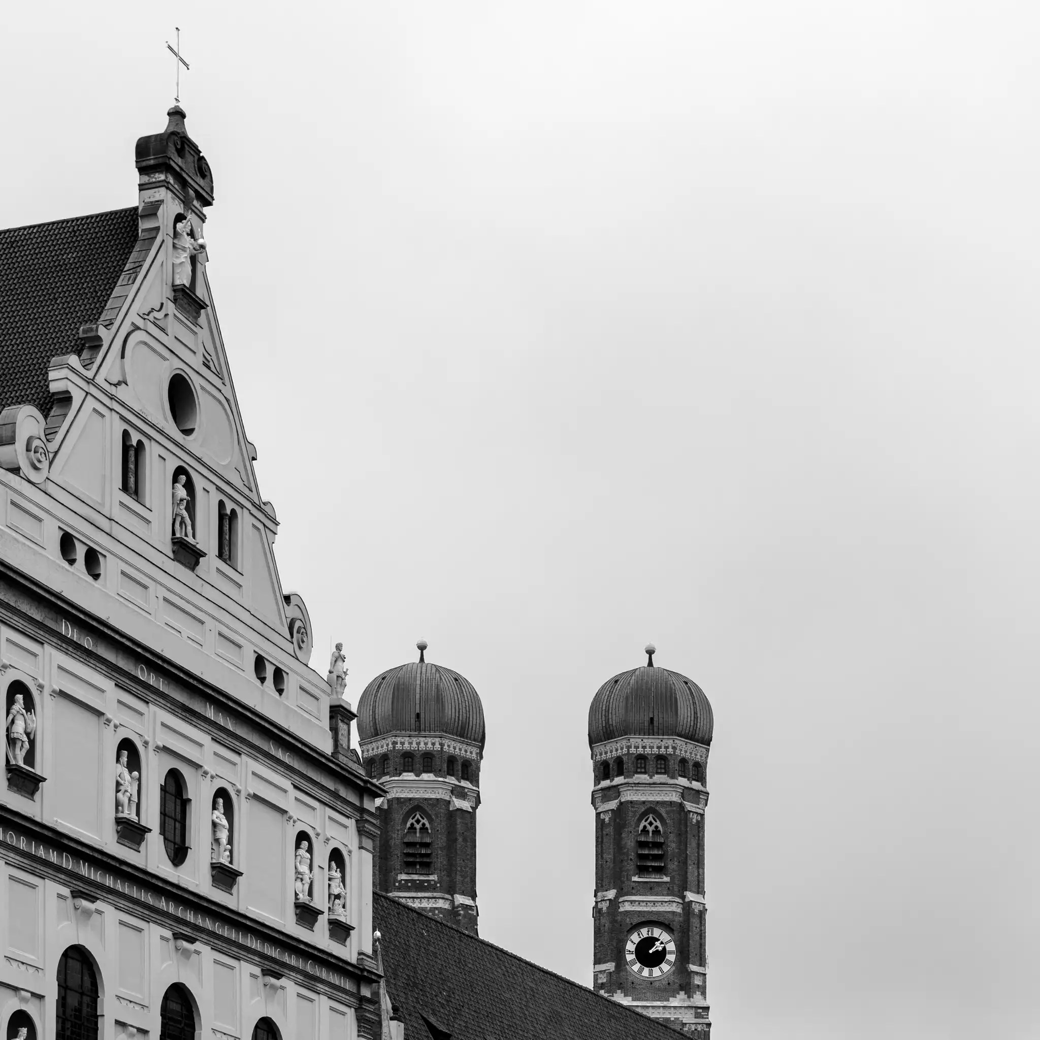 Blick auf die Zwillingstürme der Frauenkirche und ein barockes Gebäude mit Statuen in München bei bewölktem Himmel.