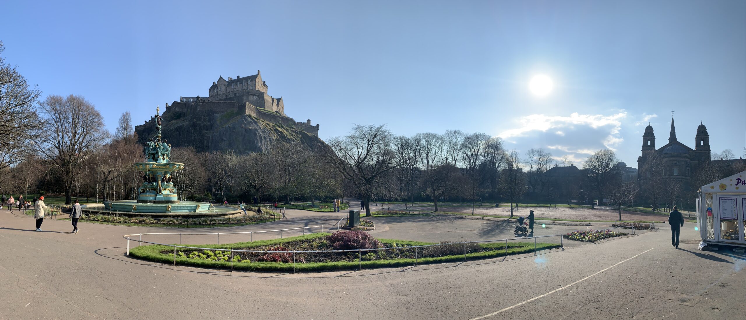 A panoramic view of Edinburgh Castle from Princes Garden, including a fountain and church, with the sun shining.