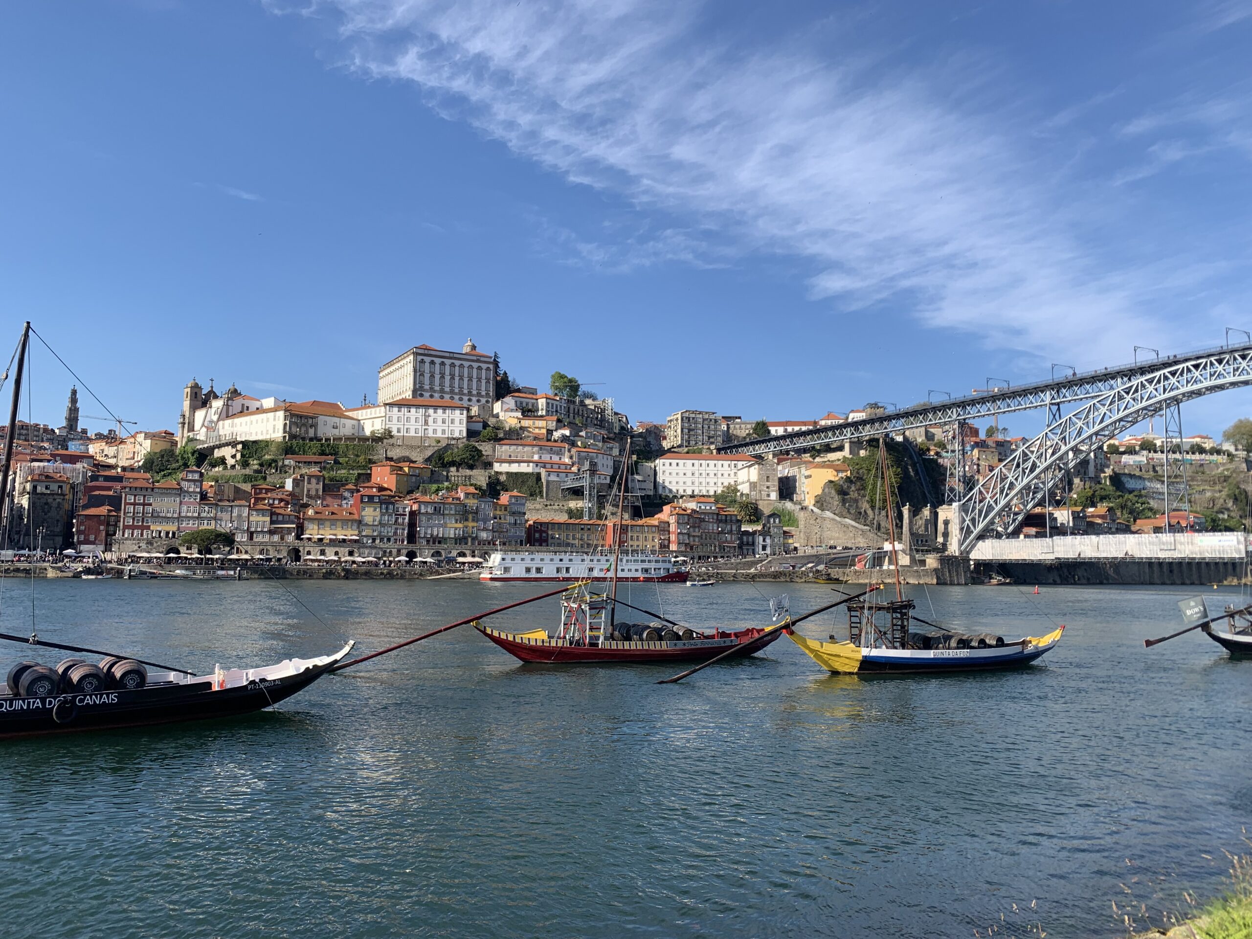ships used to carry port are docked in the river with the hills of Porto city rising behind them and a large metal bridge reaching across the river on the right