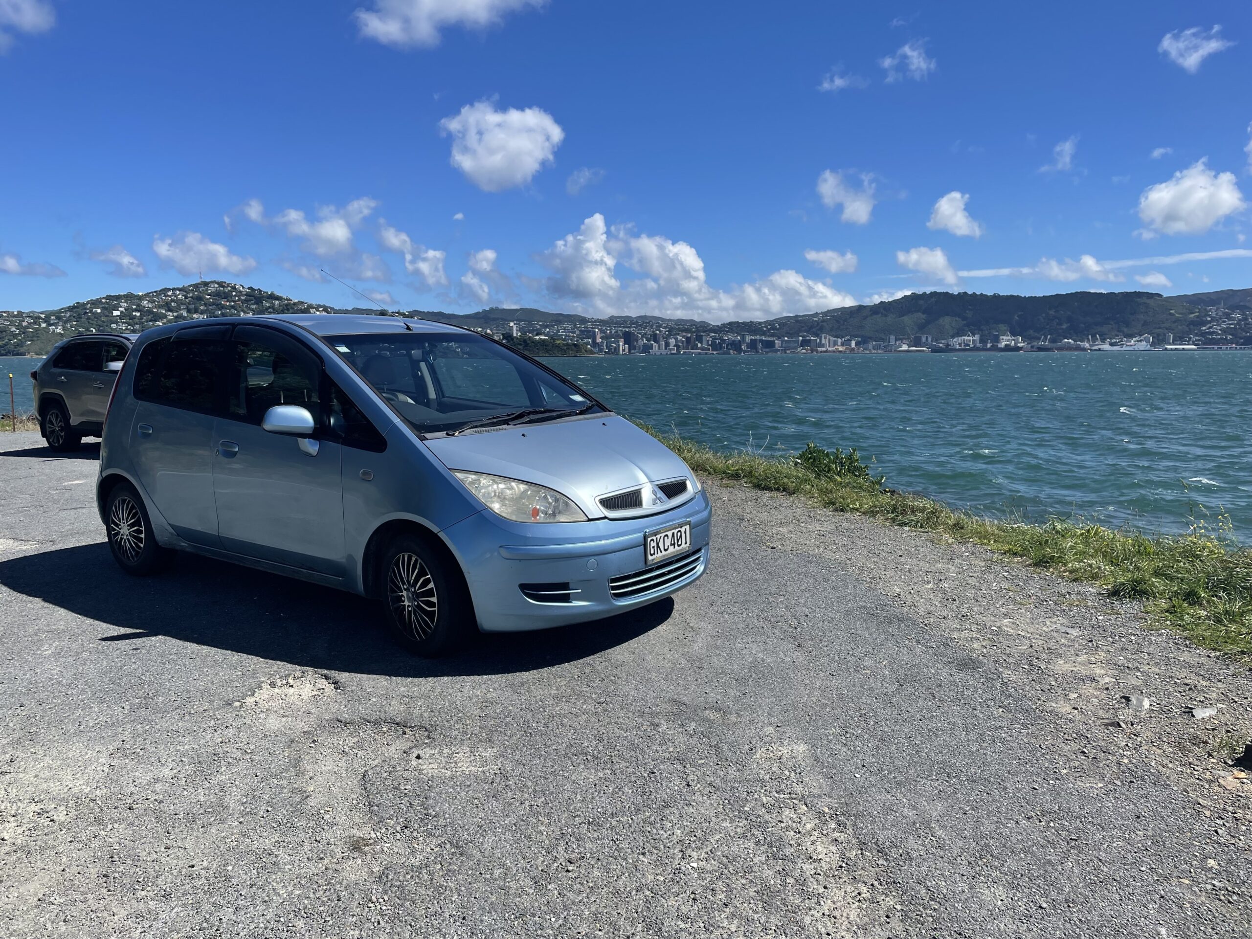 A light blue hatchback car parked on the side of a gravel road next to the sea.