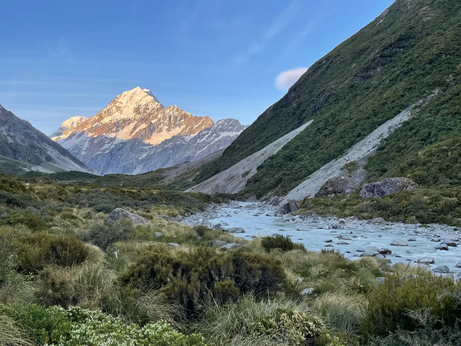 A river flows in front of the trail through brush with Mount Cook in the distance at sunset