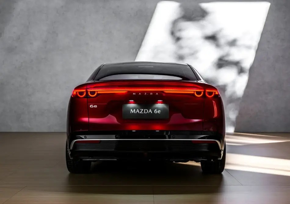 Vista trasera de un Mazda 6e berlina rojo en una sala de exposición, con las luces traseras encendidas y el emblema «MAZDA 6e» visible en la zona de la matrícula.