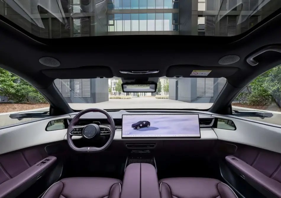 Interior de un coche moderno de lujo con una amplia pantalla táctil horizontal, volante y asientos de cuero morado; a través del parabrisas se divisa un edificio acristalado más adelante.