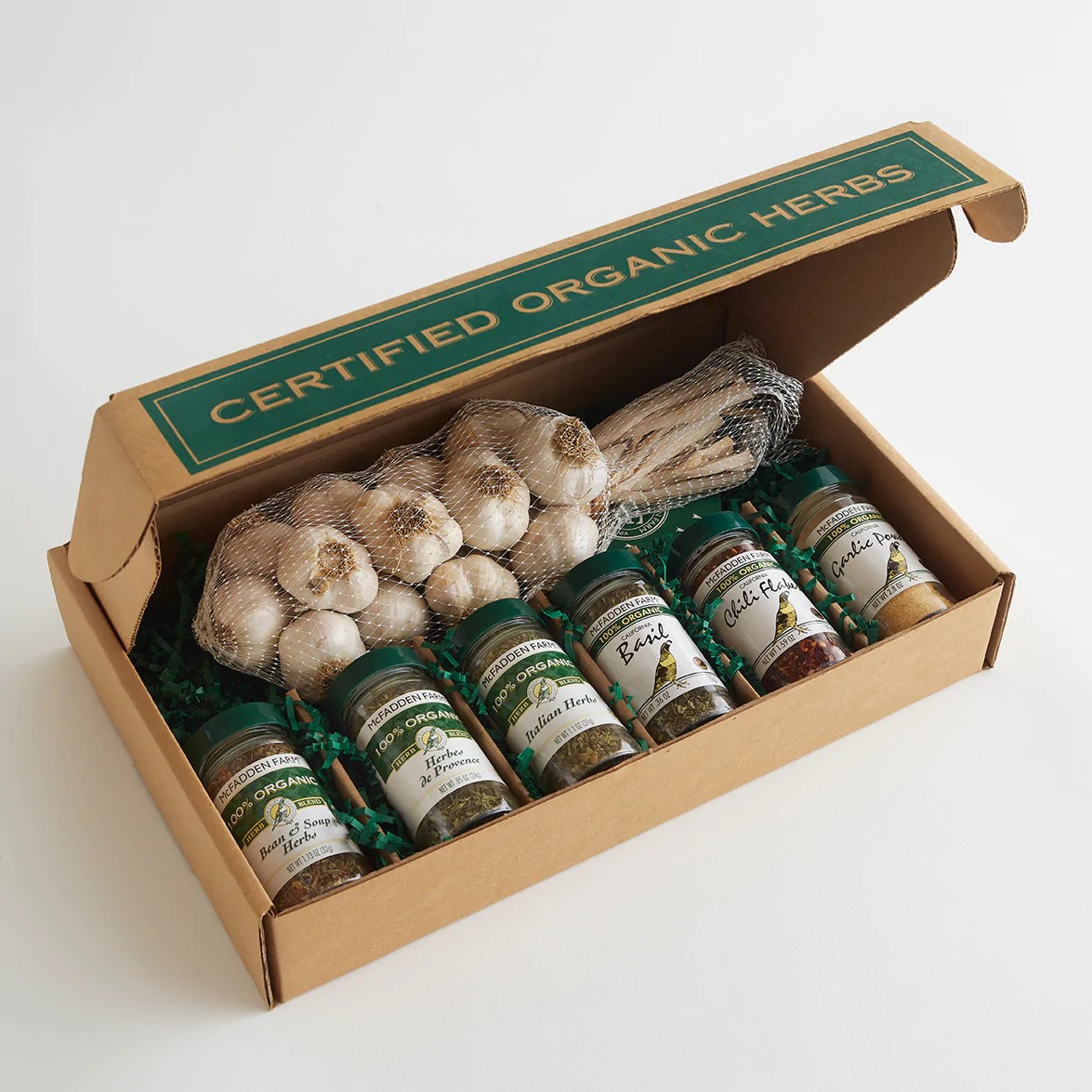An open cardboard gift box labeled “Certified Organic Herbs” contains several jars of dried herbs, a bundle of garlic bulbs in mesh, and a wrapped bundle of dried herbs arranged in green packing material.