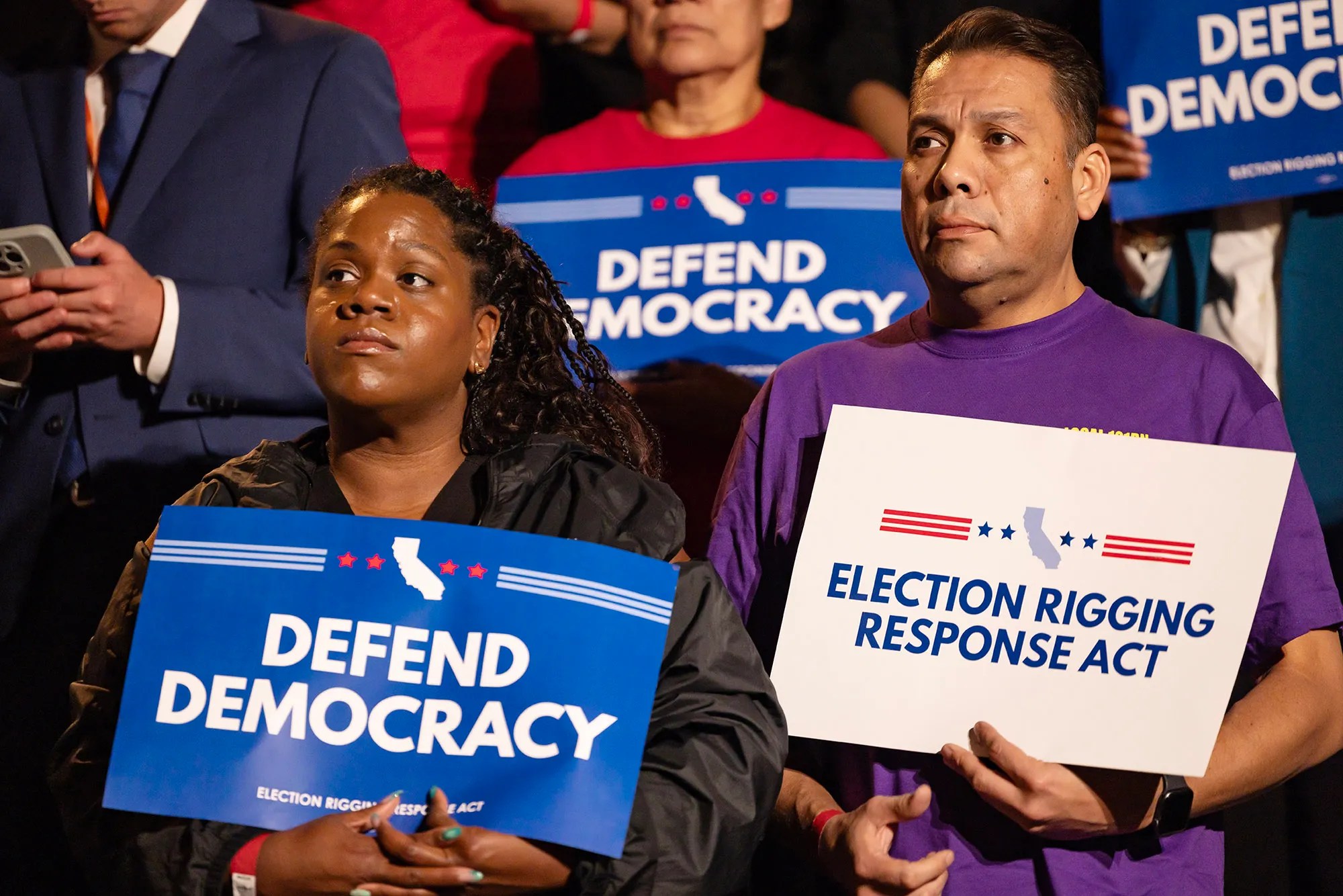 Two people hold signs that read “Defend Democracy” and “Election Rigging Response Act” at a rally, surrounded by others holding similar signs in support of California’s Proposition 50.