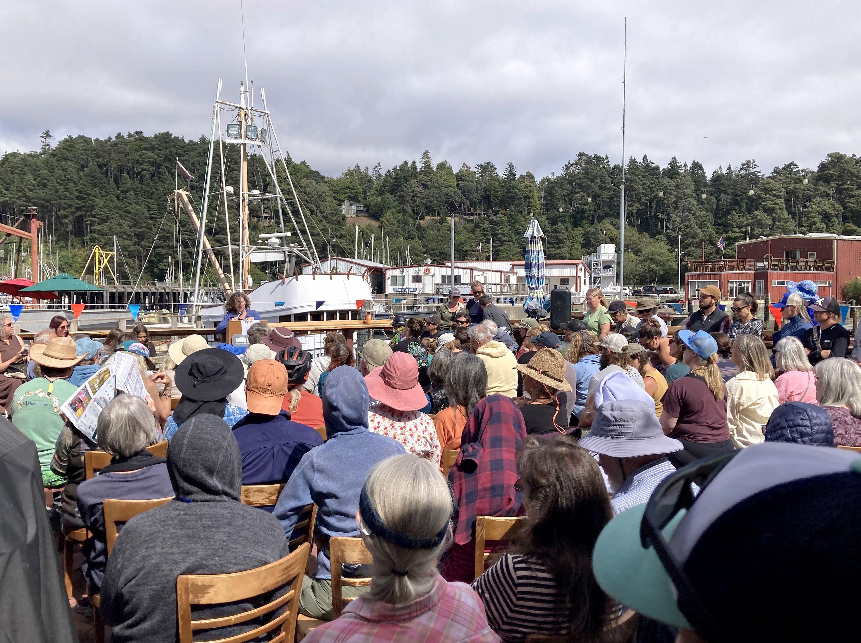 A crowd seated in chairs at an outdoor gathering near a harbor, with a large boat and forested hills in the background.