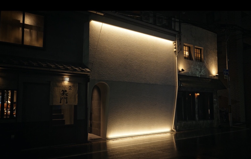 Nighttime street scene: a white stucco building with warm ground lighting along the base and an arched doorway, adjacent to a dark storefront with a hanging sign visible on the left.