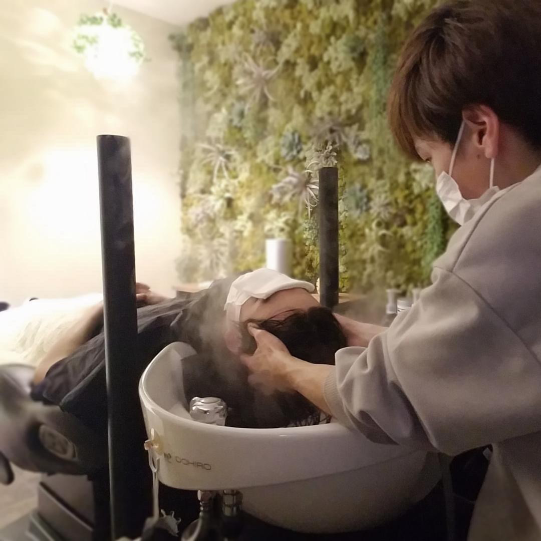Client lying back in a salon shampoo chair while a stylist wearing a mask washes their hair, steam rising from the basin in a plant-filled space.