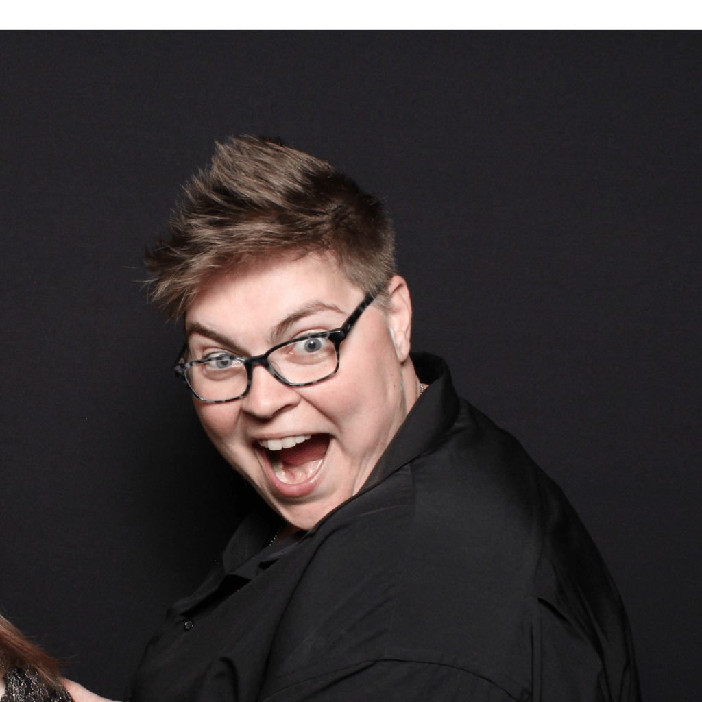 A person with short hair and glasses smiles widely against a black background, wearing a black shirt, reminiscent of an art studio setting.