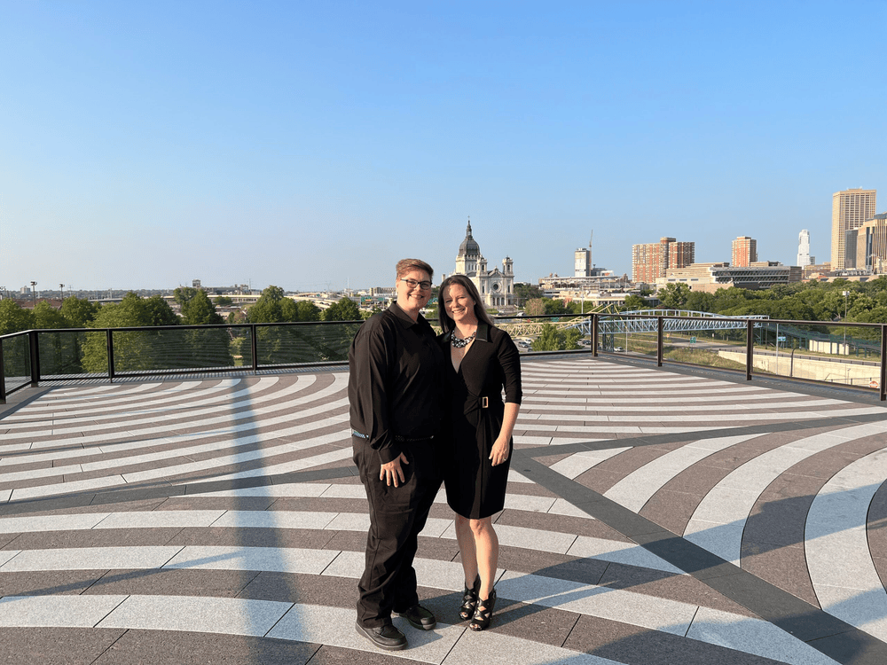 A couple stands on a patterned terrace, their black outfits contrasting with the vibrant cityscape that ignites the imagination. The clear sky frames the domed building and skyscrapers, while their poised elegance is a striking canvas against the bustling creativity of urban life.