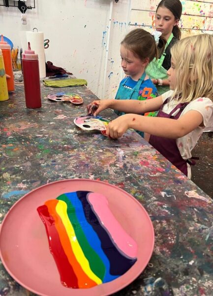 Three children in aprons are painting at a splattered table in the vibrant art studio. They hold paintbrushes and focus on a colorful palette. In the foreground, a plate with rainbow-colored paint stripes is visible, embodying the creative spirit of kids art classes.