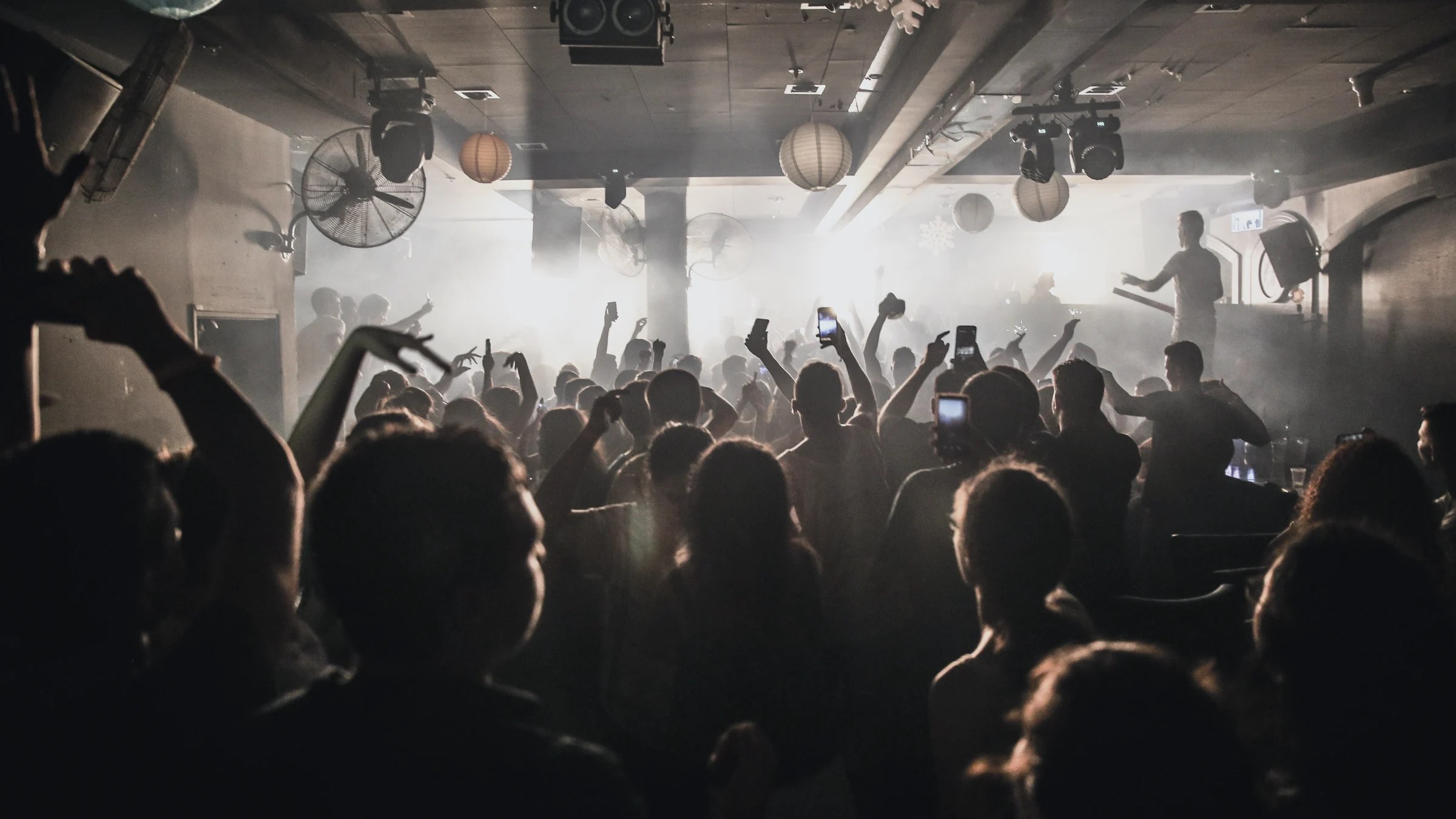 Silhouetted crowd dancing with raised hands facing bright stage lights in a nightclub.