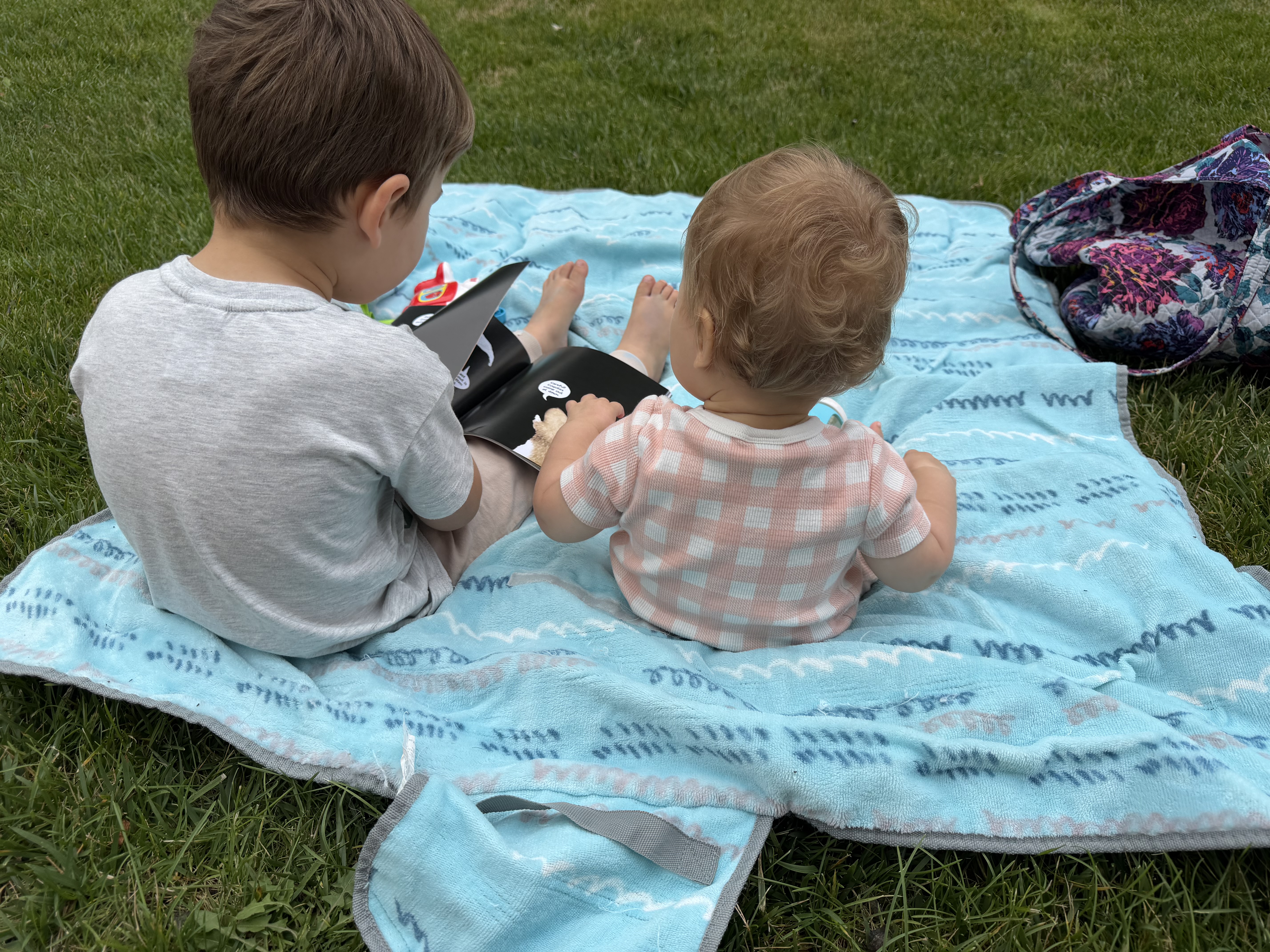 Two siblings looking at a book together on a blue blanket outdoors