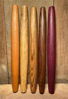 Five long narrow wooden rolling pins of varying colors with tapered ends.