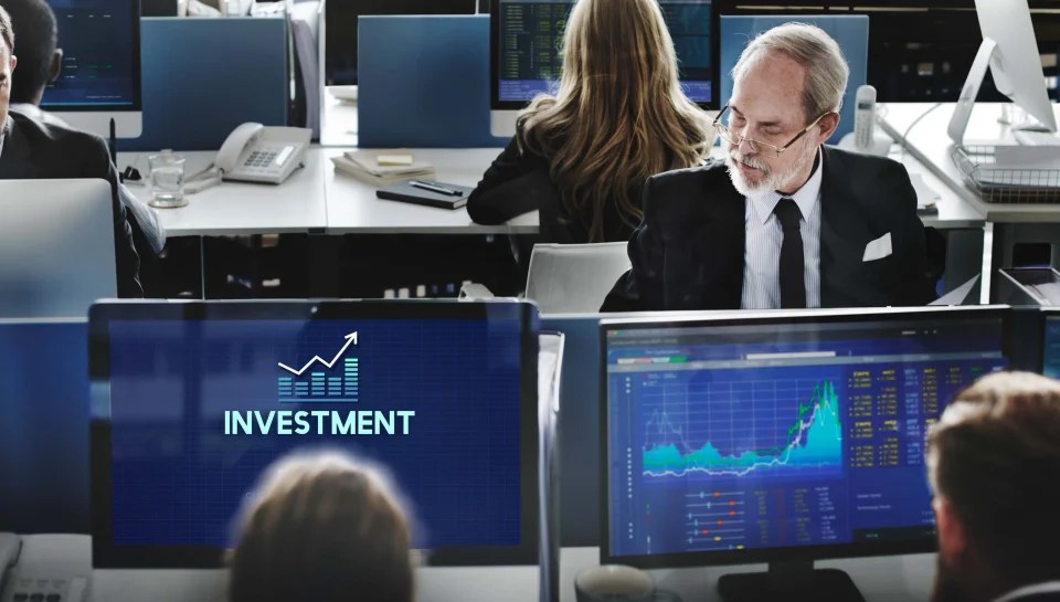 Investment professionals monitoring stock market data and financial charts on multiple trading screens in a corporate office.