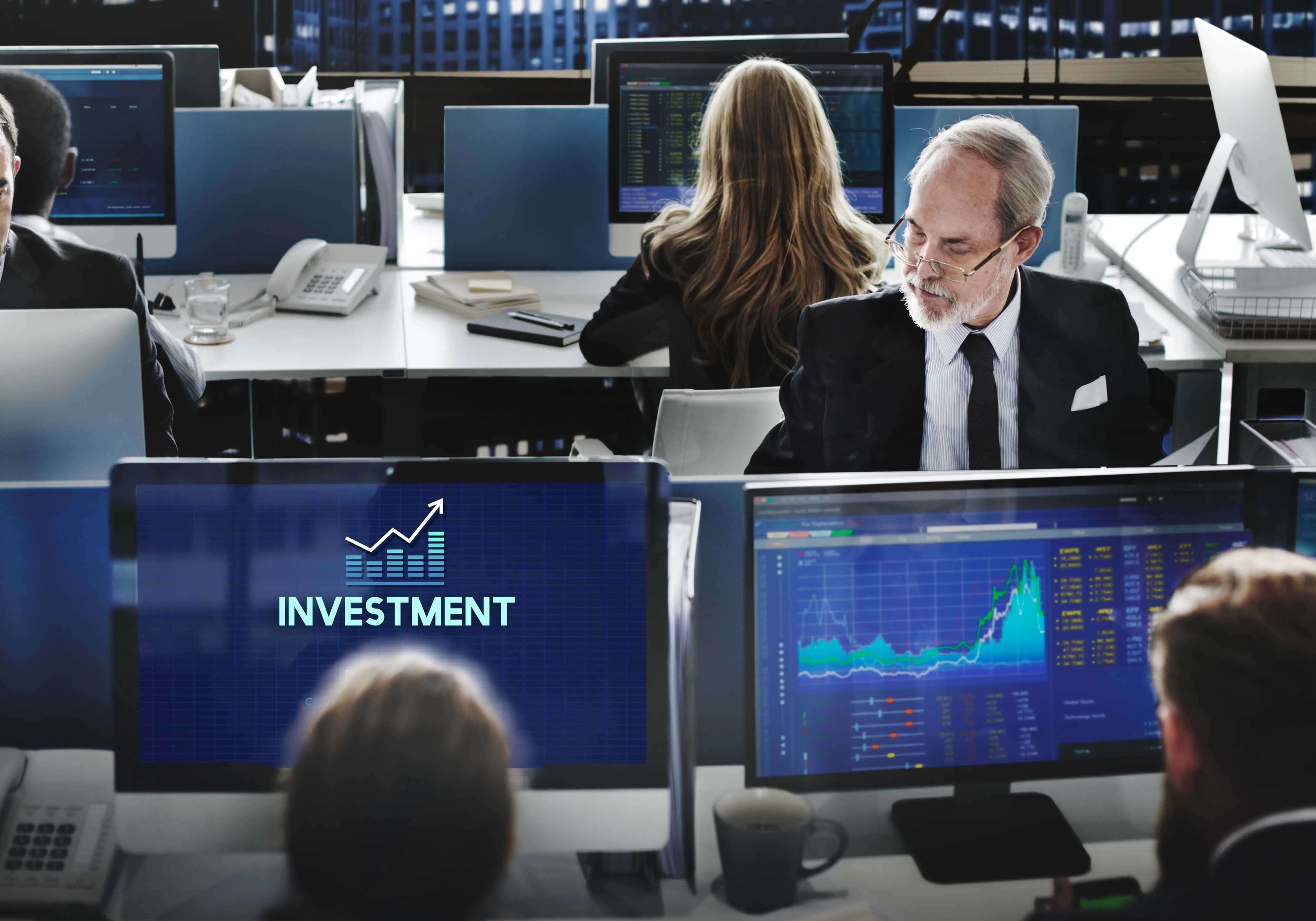 Investment professionals monitoring stock market data and financial charts on multiple trading screens in a corporate office.