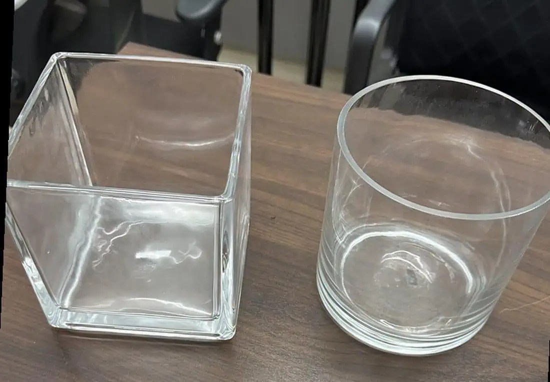 Suministro de artículos de cristalería: dos recipientes de cristal transparente colocados uno al lado del otro sobre un escritorio de madera —un jarrón cuadrado de fondo plano y un vaso cilíndrico— que representan los productos de referencia del cliente para la exposición de flores frescas