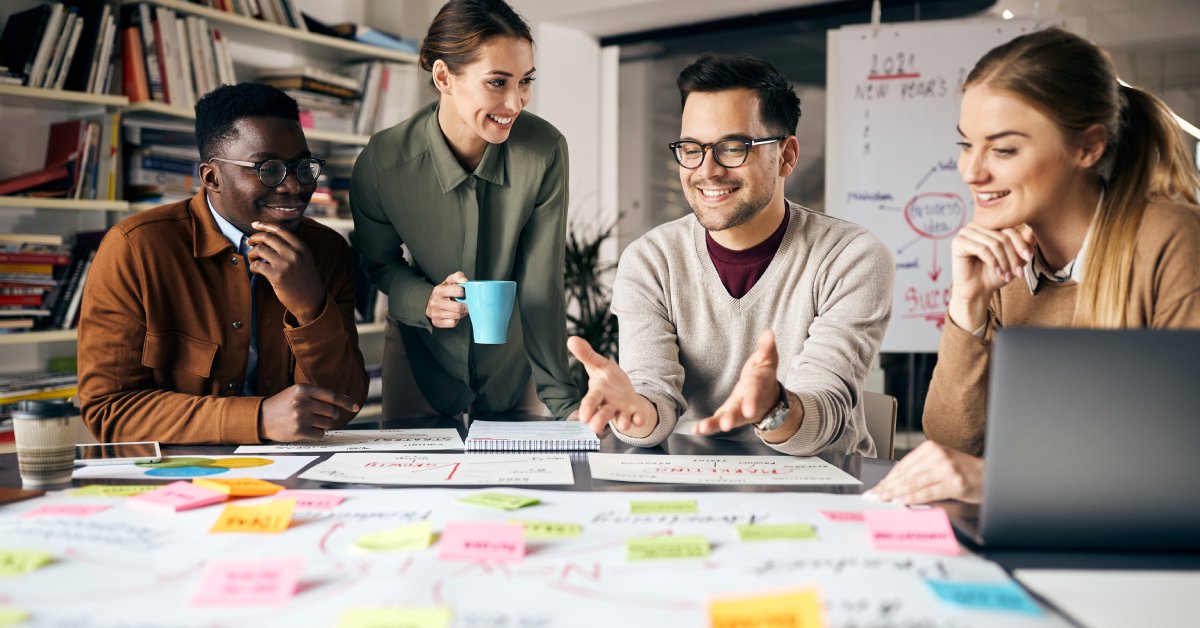 Four individuals sit at a workspace, focused on a large paper and sticky notes spread across the table.