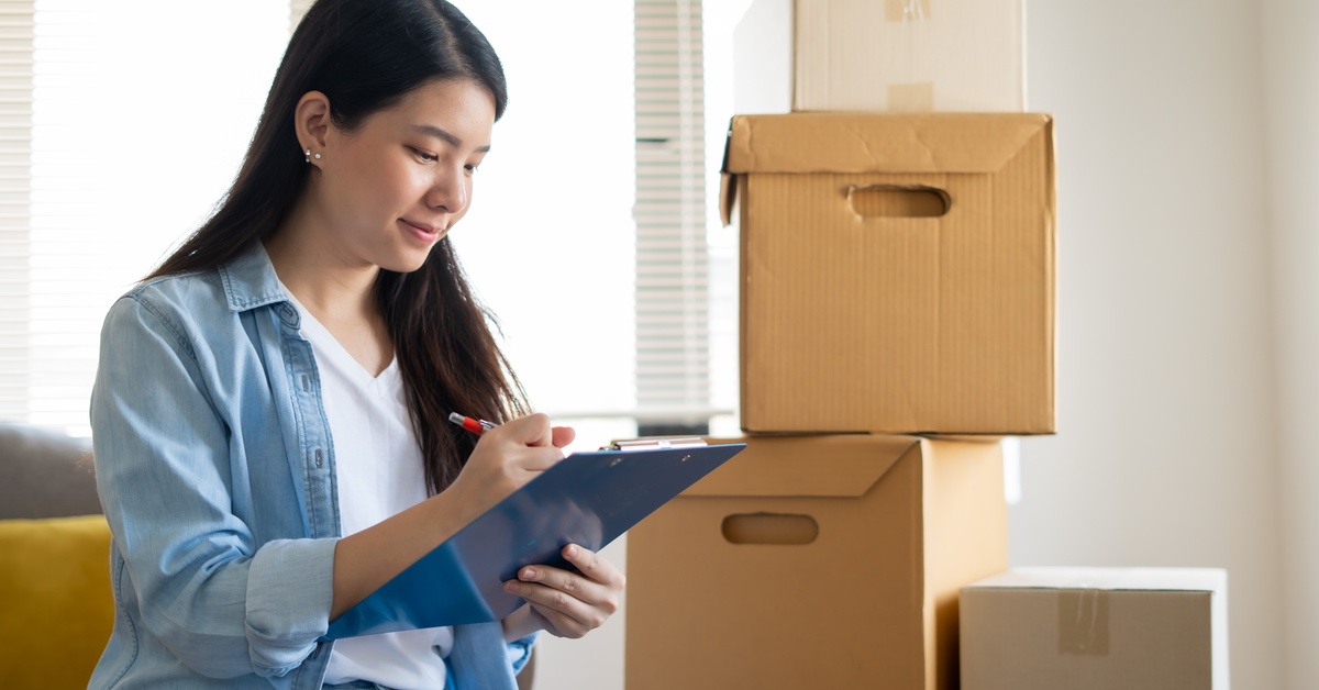 An Asian woman smiles while writing notes on a clipboard. There is a stack of cardboard boxes beside her.