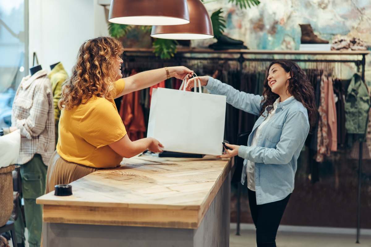 A retail employee hands a customer a large shopping bag across the checkout counter of a boutique store.