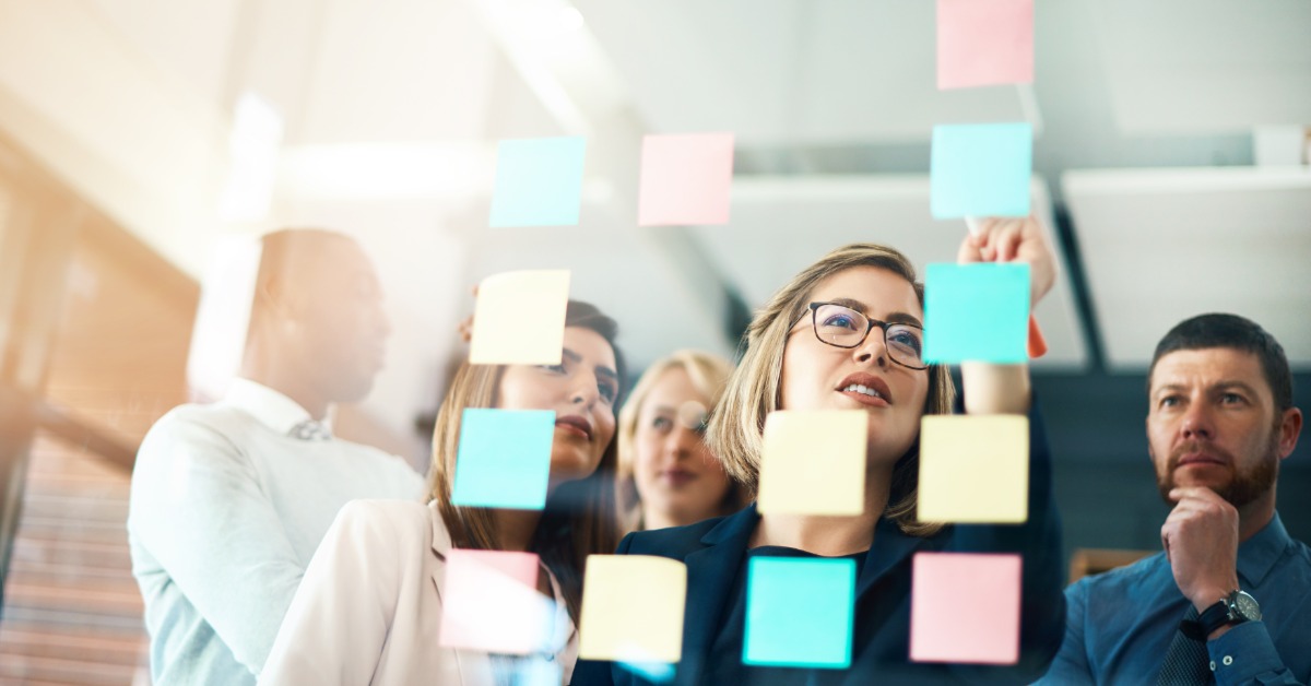 Several people, a mixture of men and women, are all looking at sticky notes stuck to a glass wall.
