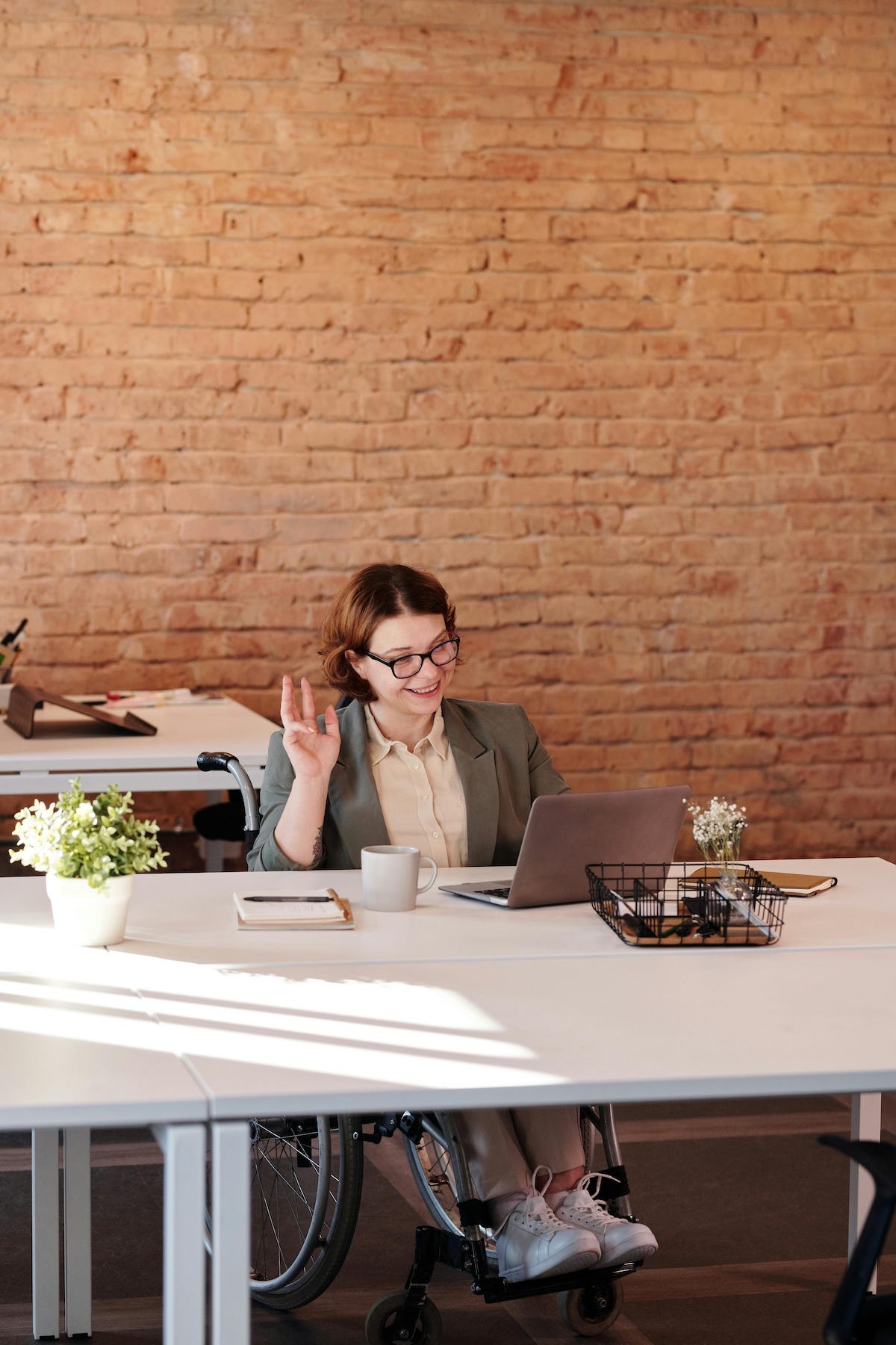 A woman wheelchair user in a blazer, working remotely. She is smiling and waving into her laptop’s webcam, letting her personality shine through.