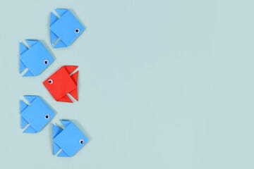 Five origami fish on a light blue background, four blue and one red. The red fish faces opposite, symbolizing individuality and contrast. This represents how God wants us to be bold for Him. Don't play it safe.