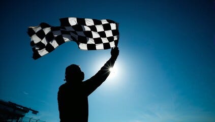 Silhouette of a person waving a checkered flag against a bright blue sky with the sun shining behind. This represents the things that might motivate us. What motivated you?