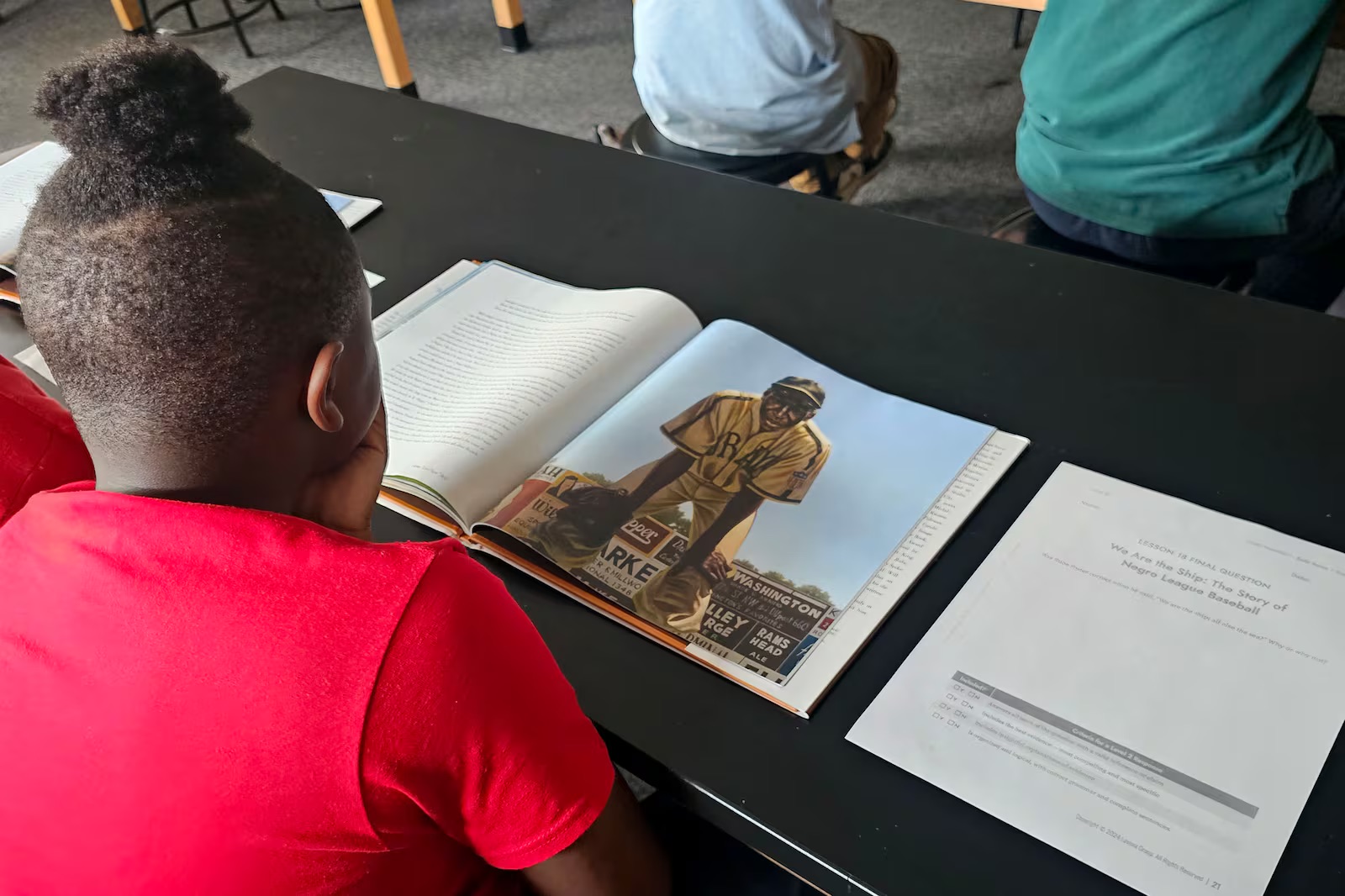 Photo looks over the shoulder of a young student who is reading a book with a worksheet nearby.