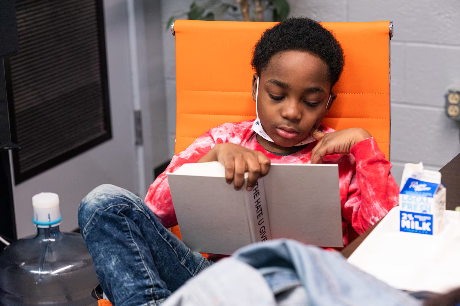 A young student lounges in a classroom chair reading the book "The Hate U Give."