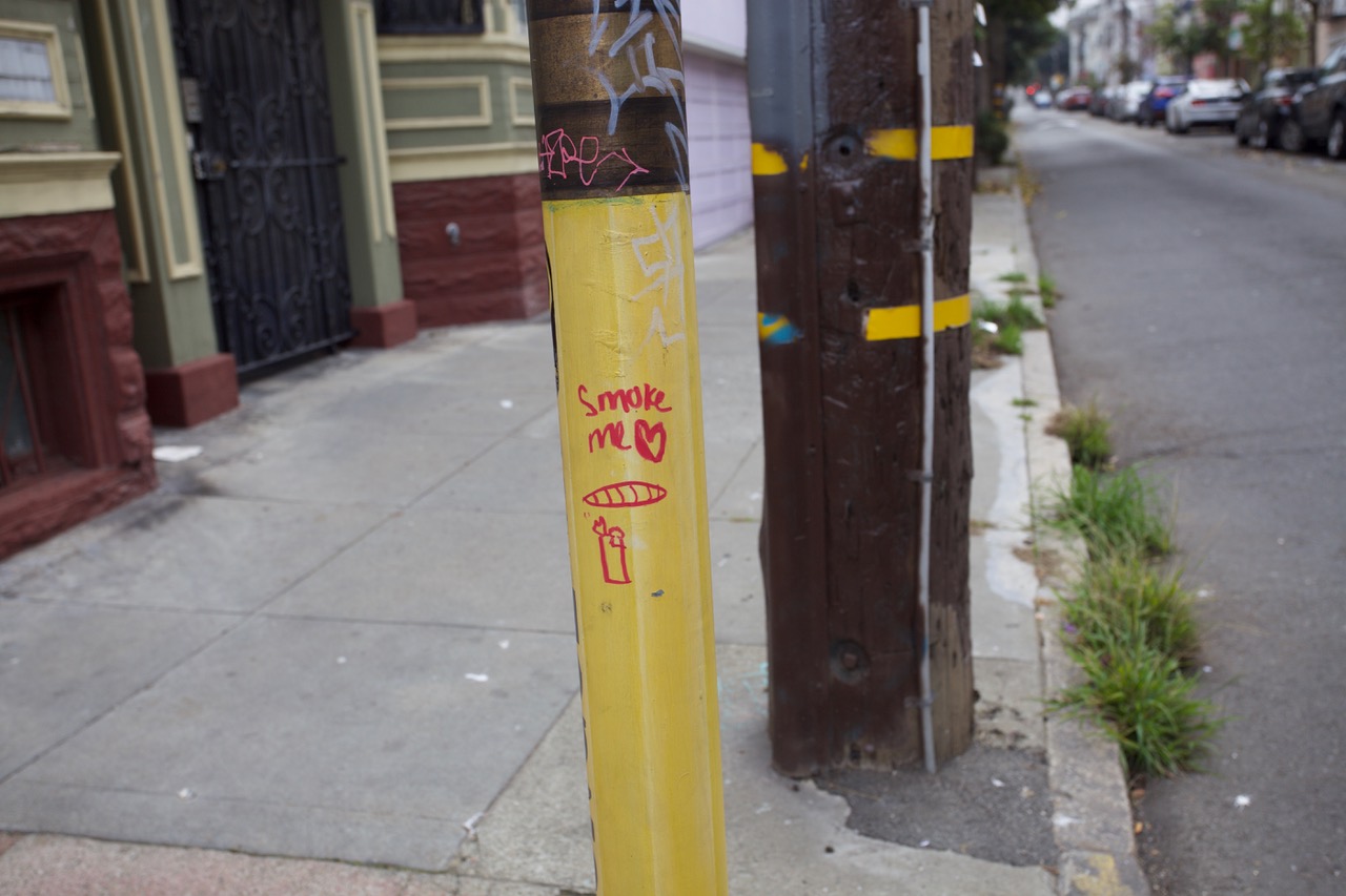 A pole with some yellow paint and writing on it that depicts a pot cigarette and the words smoke me.