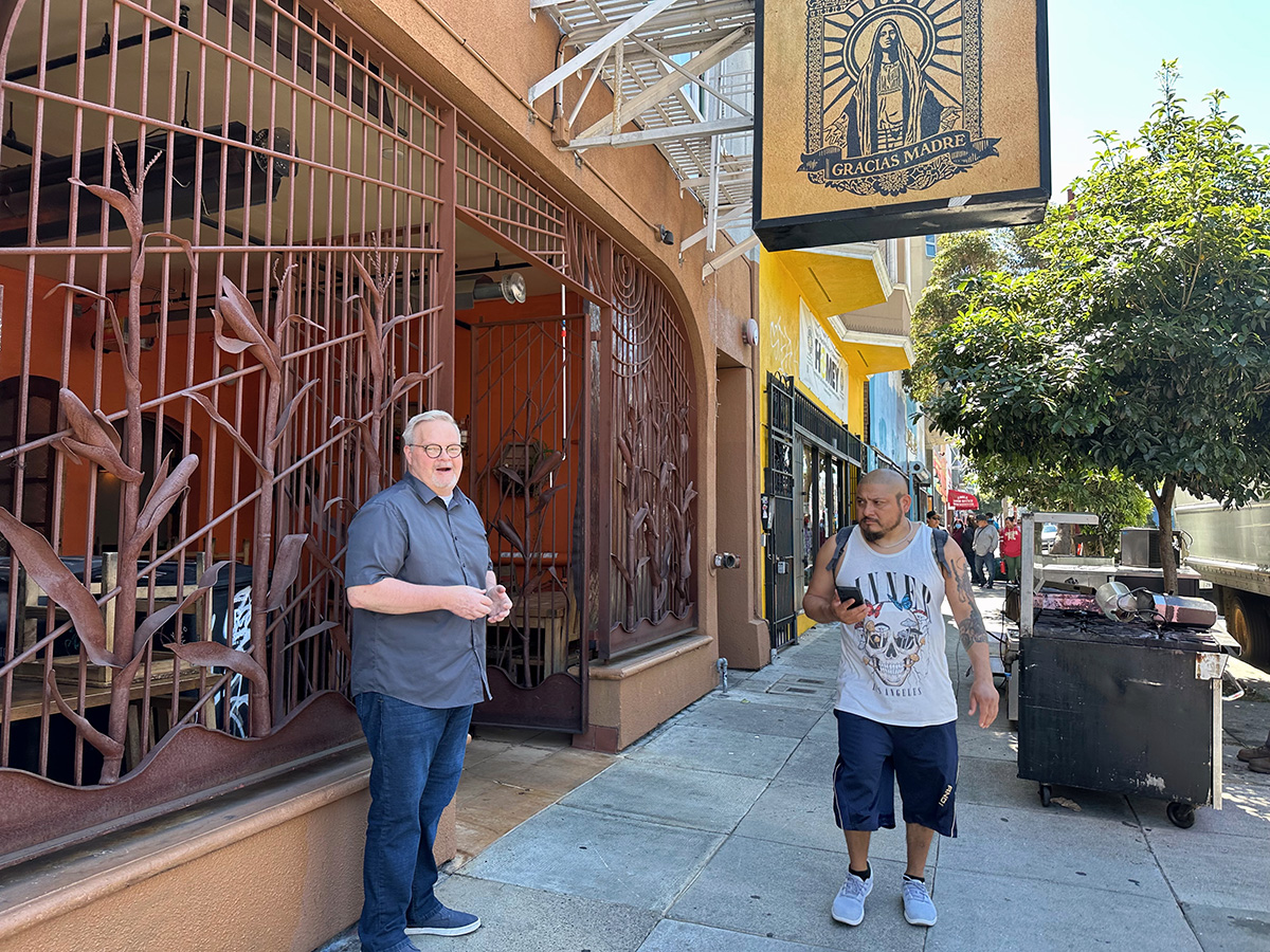 A man standing in front of a restaurant and another man walking by.