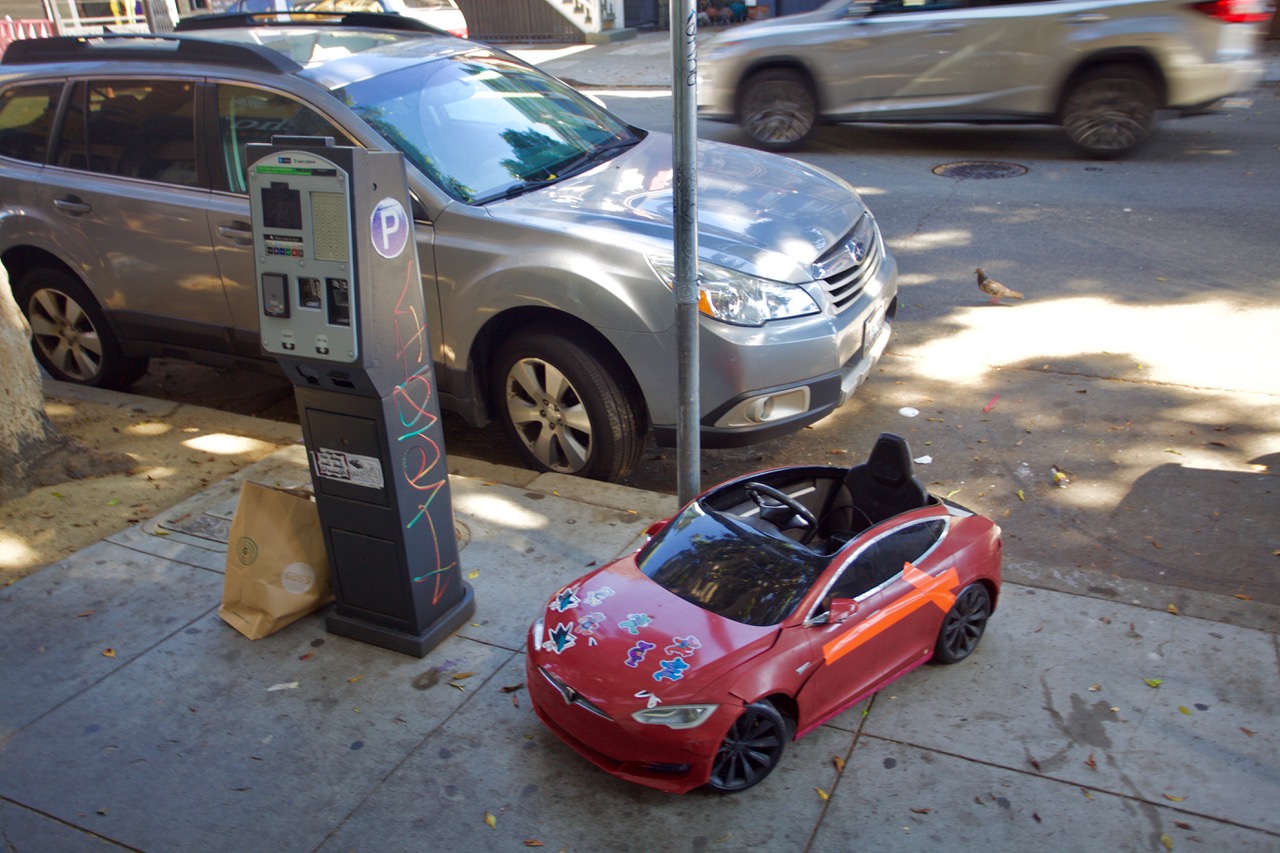 A parked car and then a toy-sized car, red, pulled up on the sidewalk.