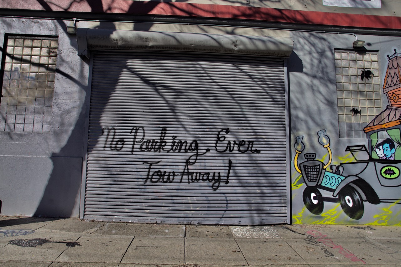 A shuttered storefront with graffiti reading "no parking ever. tow away" and colorful street art on the wall.
