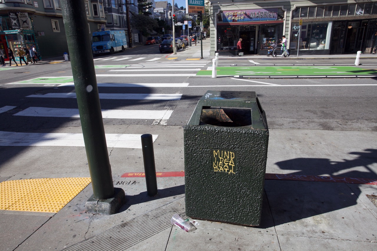 Green trash bin on a city sidewalk near a crosswalk and bike lane. Spray paint on the bin reads MIND IDEA NEAT DATA