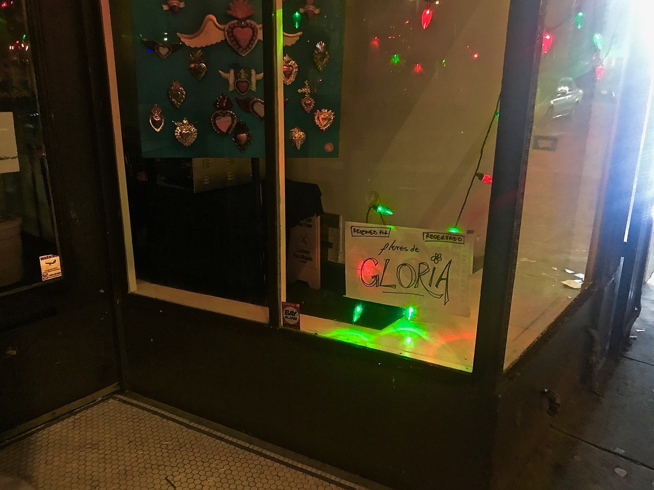 A storefront at night adorned with colorful string lights and a handwritten sign reading "flores de gloria" placed by the window.