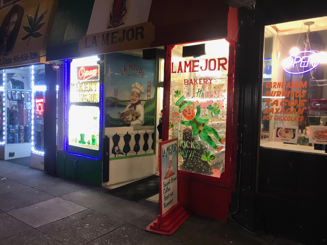 A colorful storefront at night displaying signs for "la mejor bakery" with illuminated advertisements for various food items.