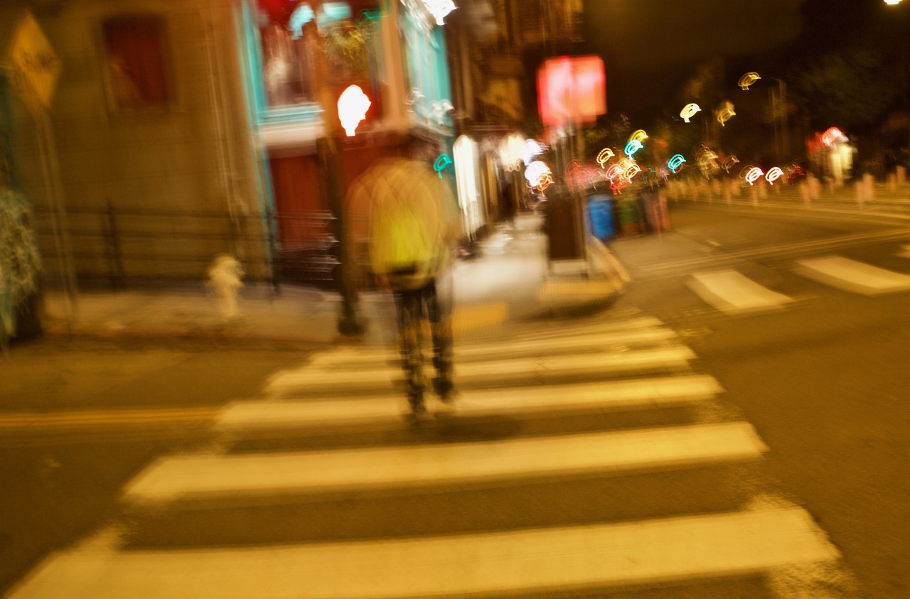 Blurry image of a person crossing the street at night with city lights in the background.
