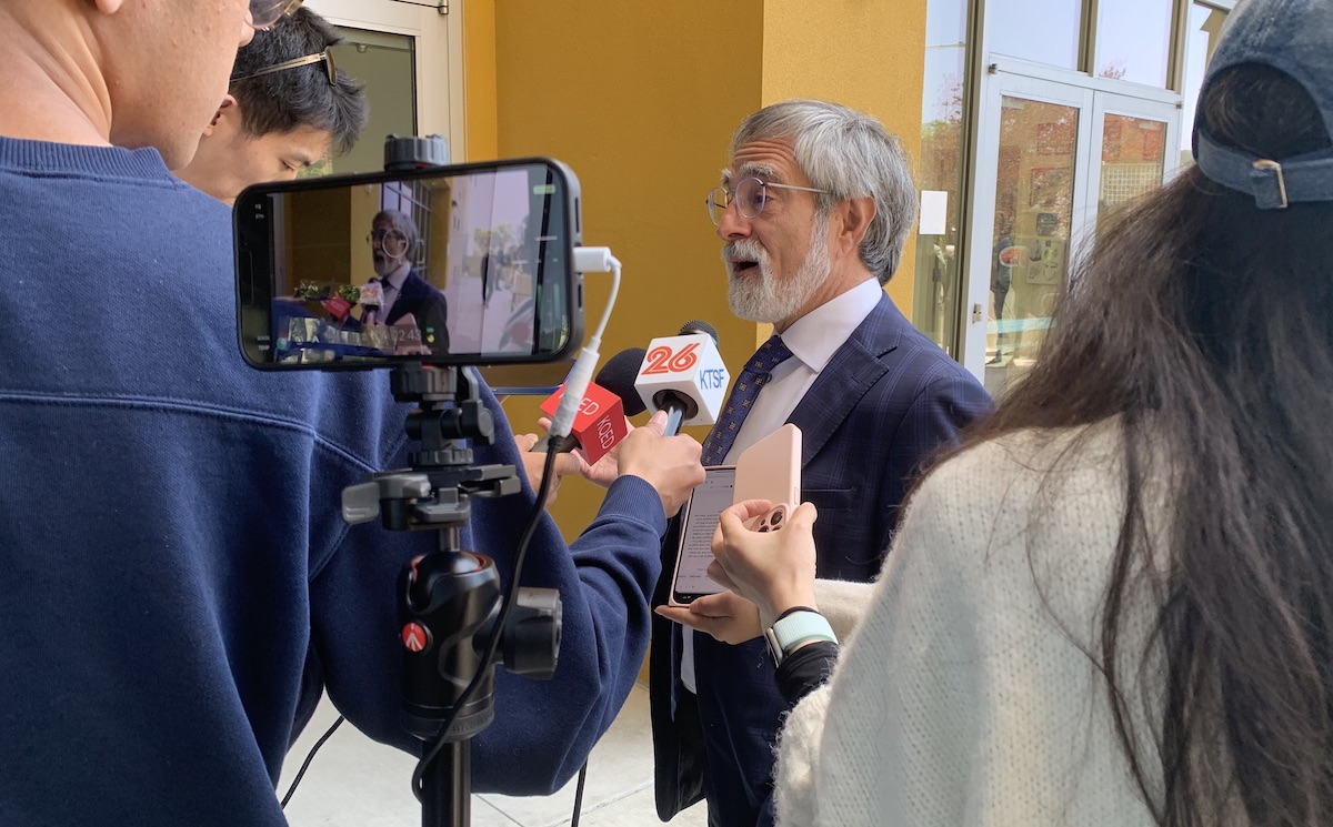 A man in a suit speaks to multiple reporters holding microphones and recording devices in front of a building. A camera on a tripod is also capturing the scene, as Peskin addresses the media with poised confidence.