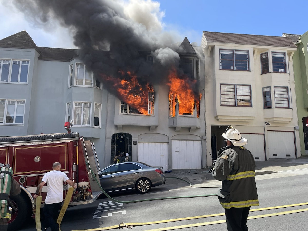 Firefighters respond to a house fire with flames and heavy smoke coming from the windows of a two-story building. A fire truck and hose are in use, and Fire Chief Williams is on scene, meticulously assessing the situation.