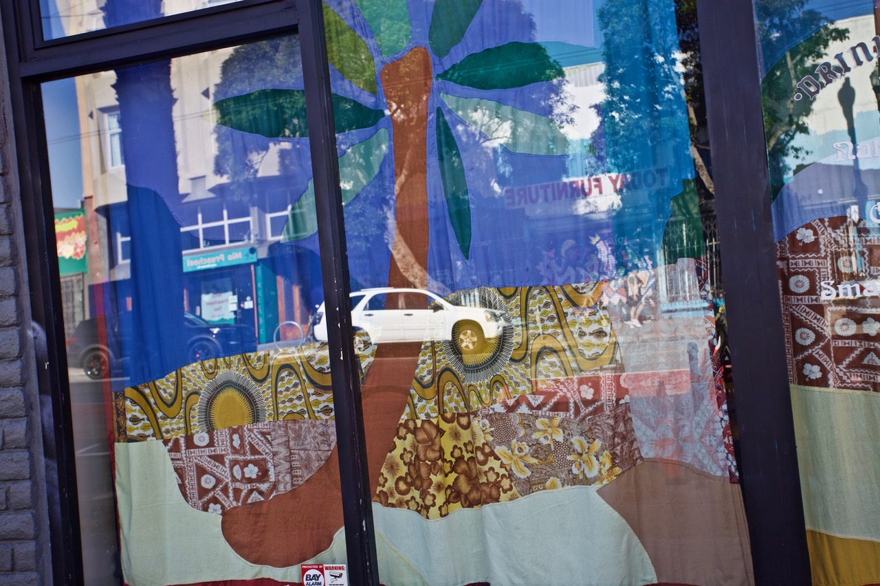 A storefront window decorated with a fabric tapestry depicting a palm tree. A reflection shows a white vehicle and street with buildings in the background.