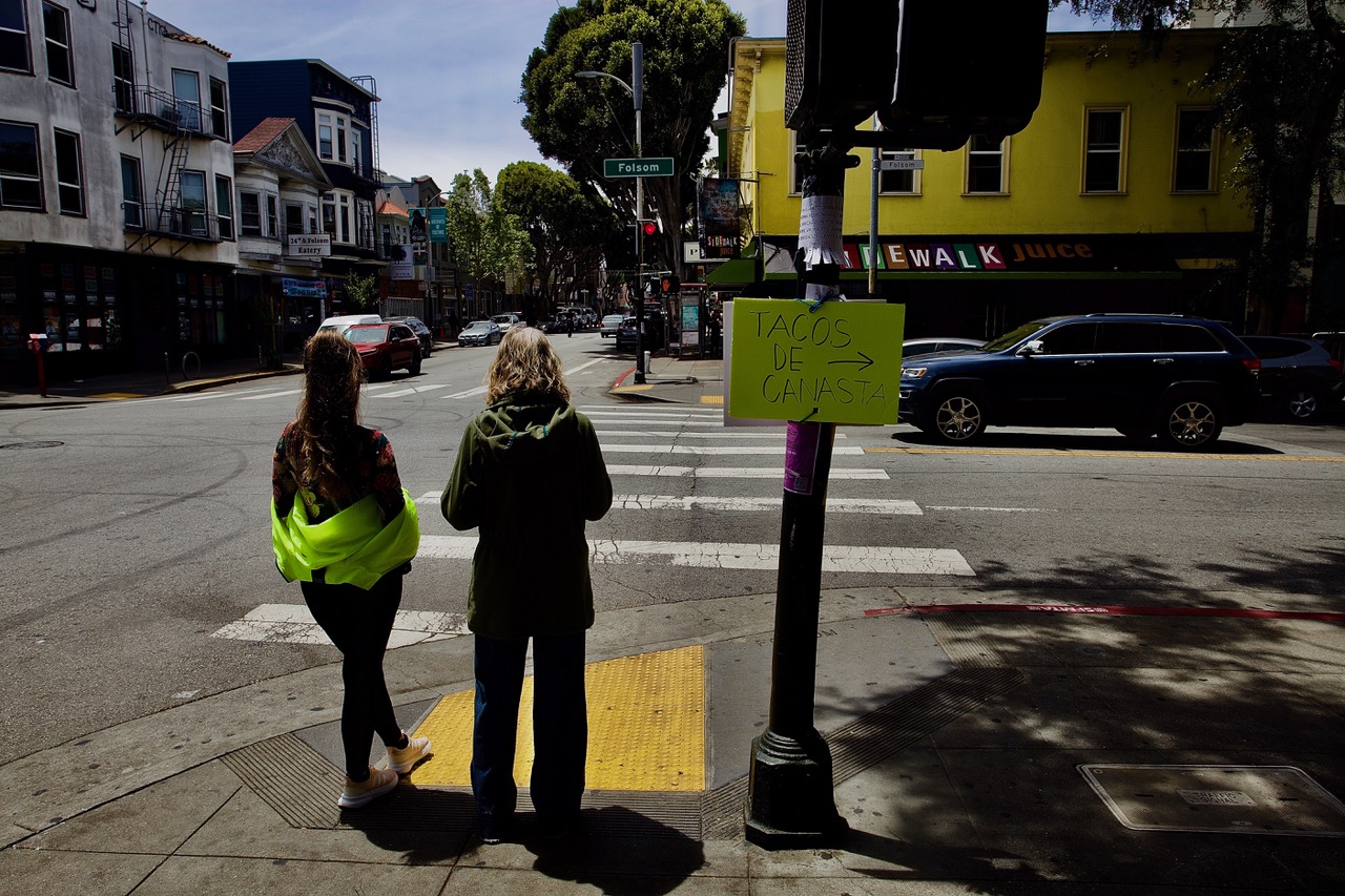 Two people stand at a street corner next to a pole with a yellow sign that reads "Tacos de Guiso" with an arrow pointing right. Buildings and parked cars are visible in the background.