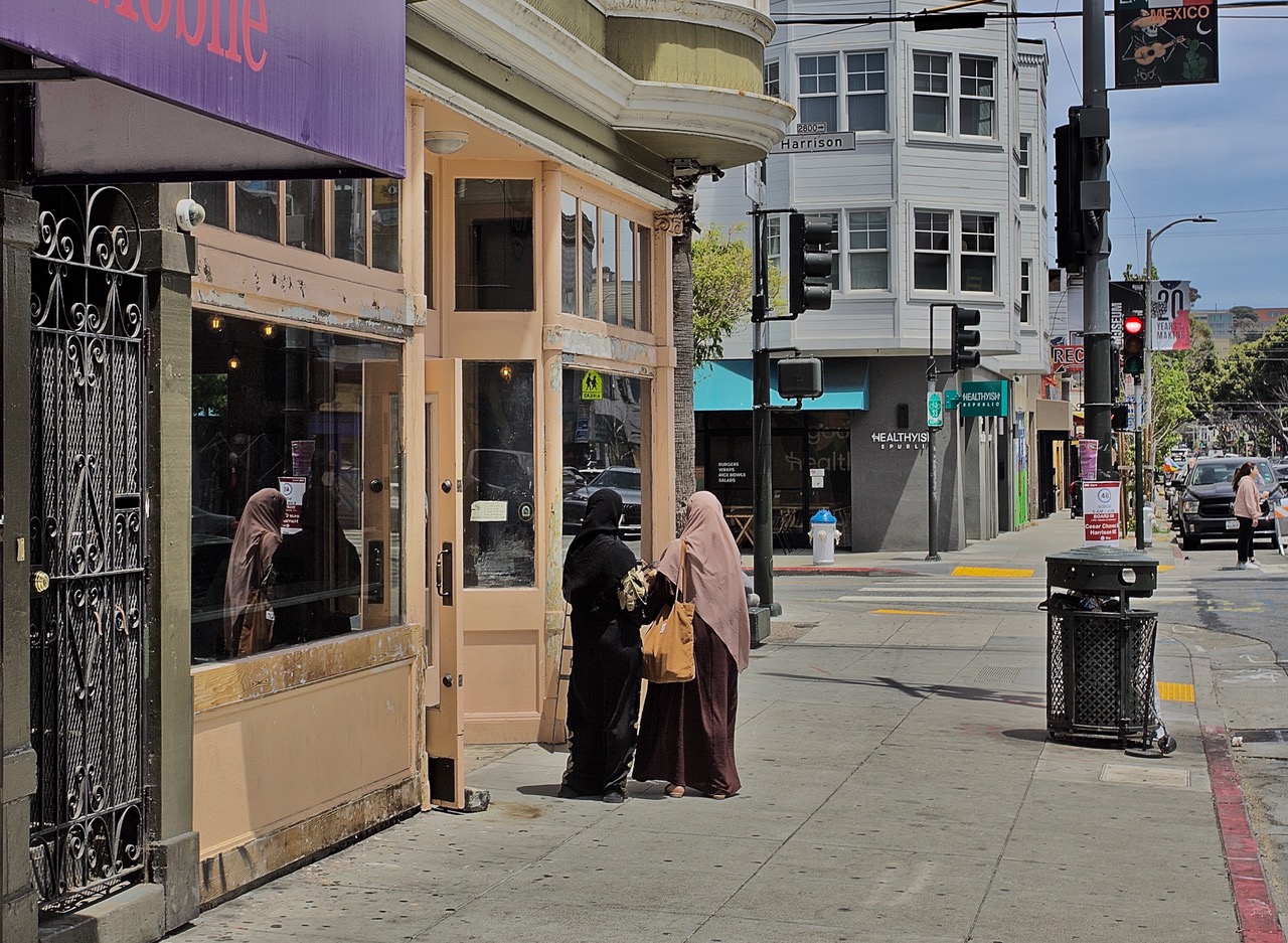Two people wearing long garments stand outside a storefront on a city sidewalk. A person is visible inside the store. There are traffic lights, buildings, and a street in the background.