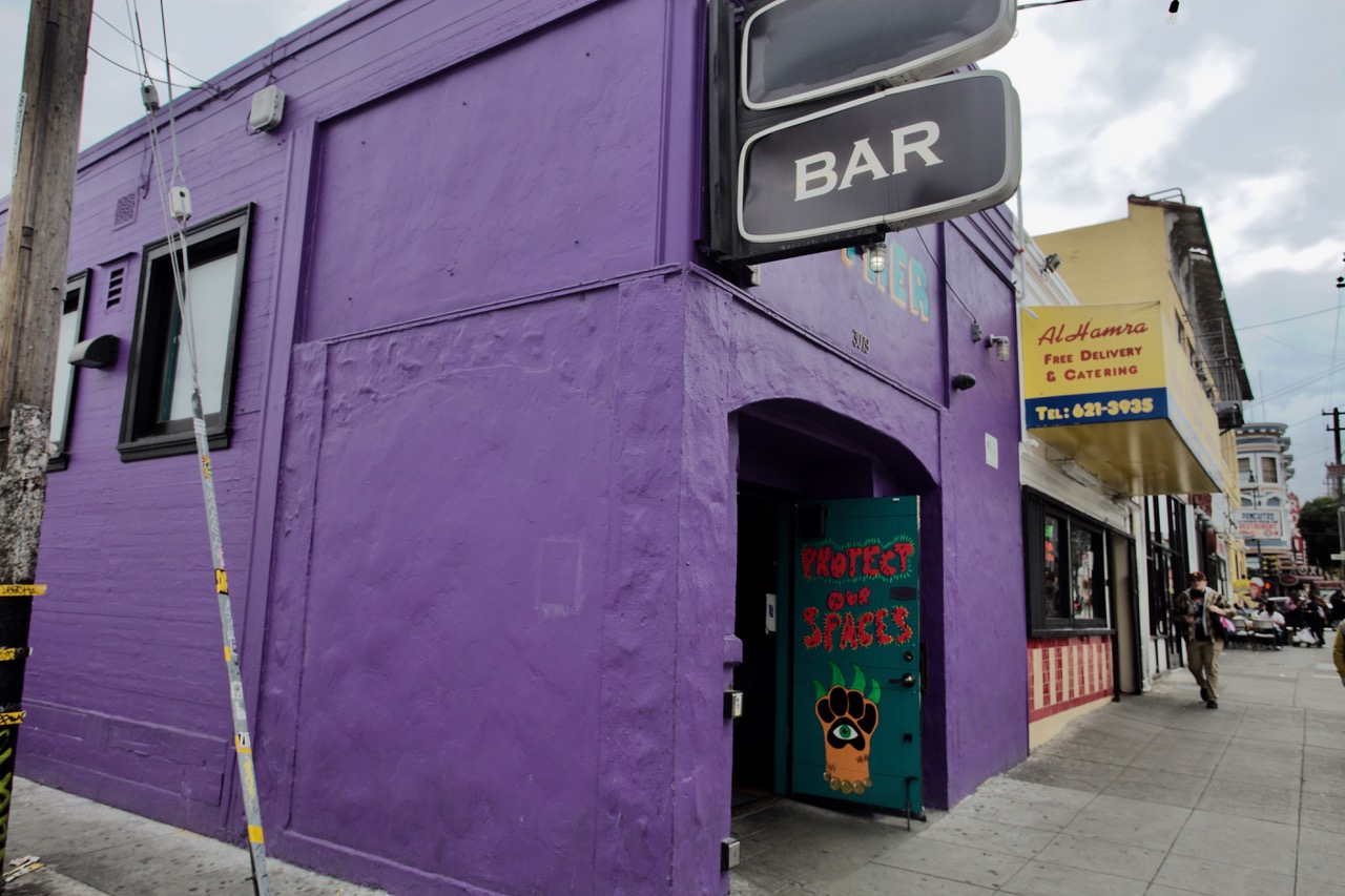A purple building with a sign reading "BAR" above the entrance. The door displays the words "Protect Our Spaces" with a paw print. Adjacent is a yellow building with a food delivery sign.