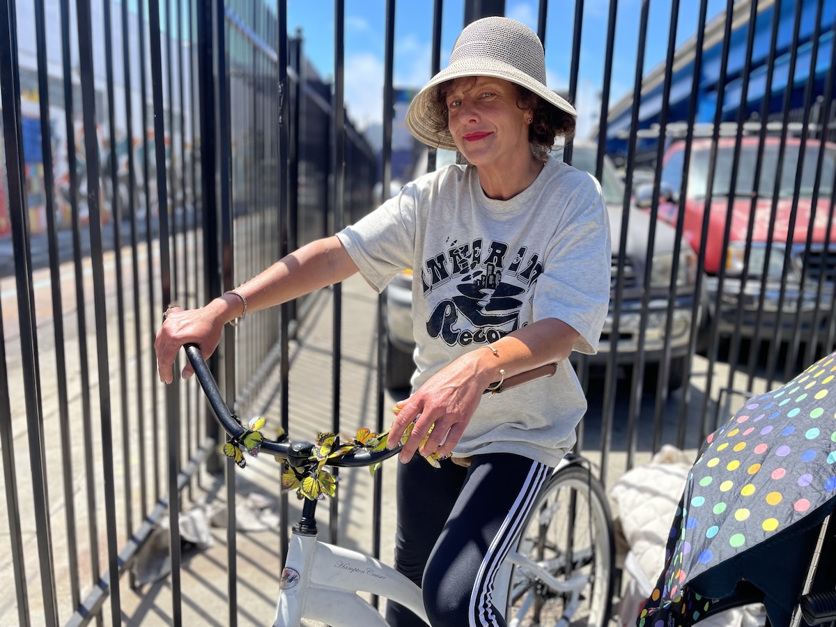 A woman wearing a hat and a t-shirt sits on a white bicycle next to a fence. She has a colorful stroller in front of her.
