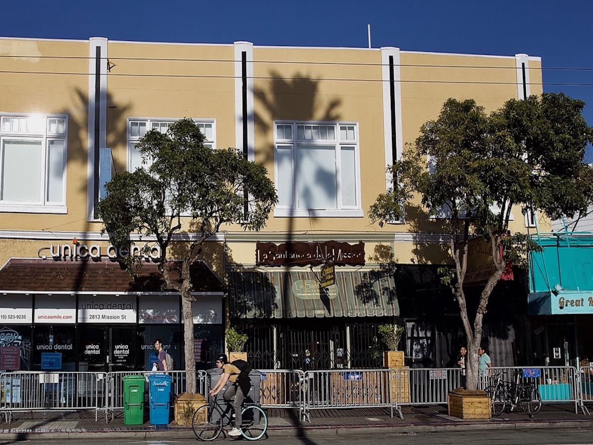 A person rides a bicycle past a yellow building with trees, various business signs, and a shadow of a palm tree cast on the facade. There are barriers and bins in front of the building.