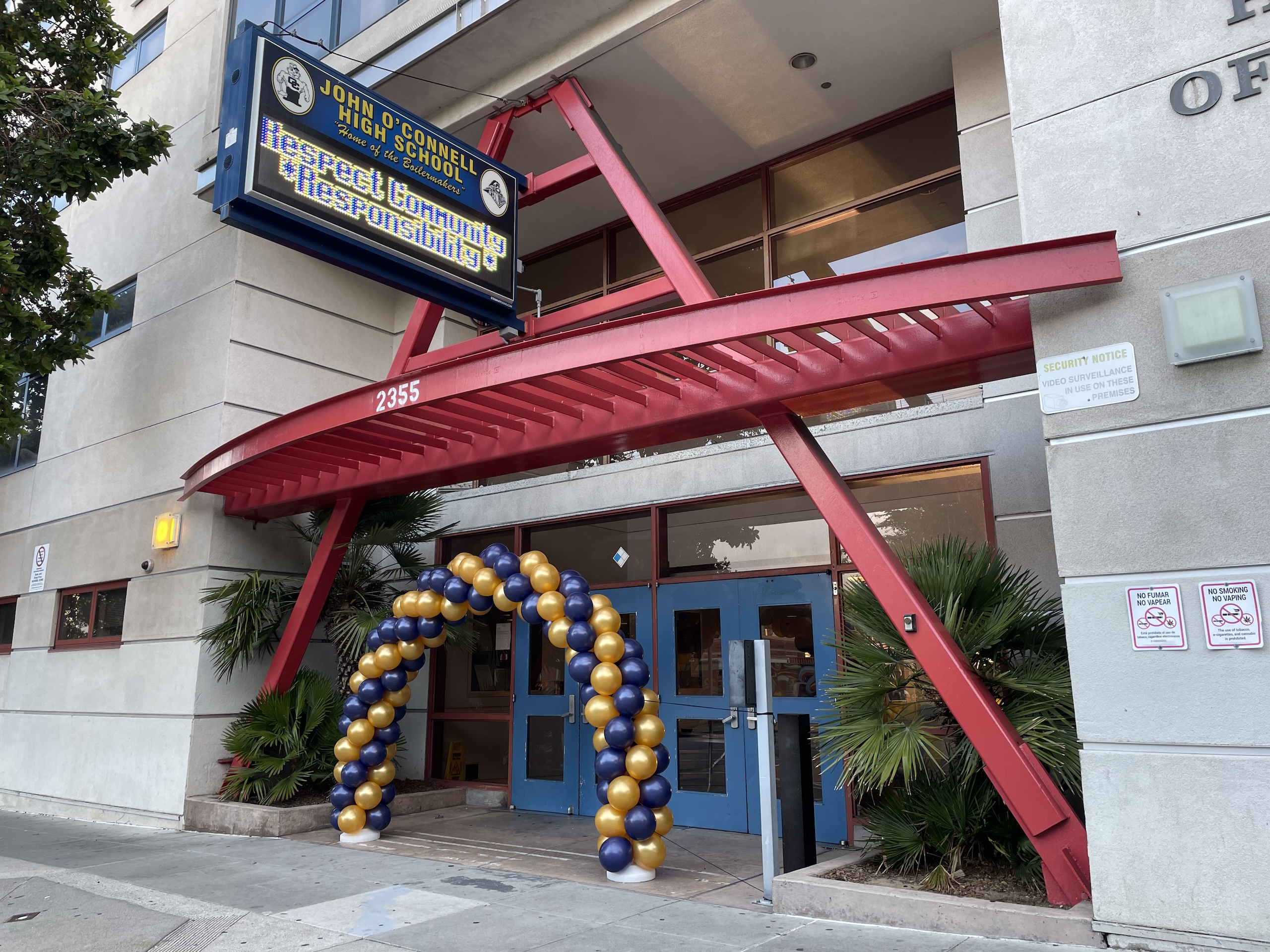 Entrance of John O'Connell High School with a banner emphasizing respect, community, and responsibility. Balloon arch made of gold and dark blue balloons decorates the doorway.
