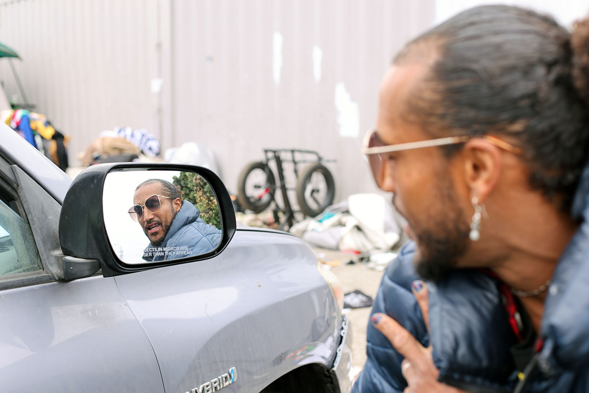 A person is reflected in the side mirror of a vehicle, looking at themselves. They wear sunglasses and have their hair tied back in a neat sweep. The background shows miscellaneous items and a bicycle.